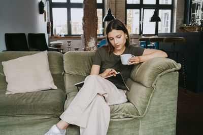 Young entrepreneur reading book having coffee while sitting on sofa in studio