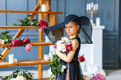 Portrait of girl wearing hat while standing with flowers against wooden steps at home