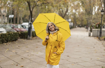 Happy senior woman with yellow umbrella standing on footpath