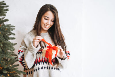 Young woman with dark long hair in cozy sweater with gift box in room with christmas tree at home