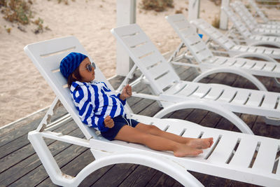 Boy child on the beach with sand. lying on a chaise longue in a blue striped jacket and sunglasses 