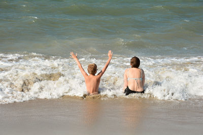 Two children sit full of joy on the sandy beach in the middle of the waves by the sea,  netherlands