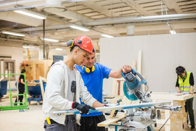 Teenage carpenter using level while standing by instructor with electric saw at illuminated workshop