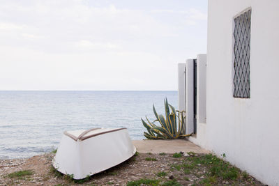 Upside down white boat on the beach, next to a white house and a green plant. calm sea. altea, spain