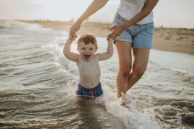 Midsection of mother with cheerful baby son enjoying in water at beach on sunny day during sunset