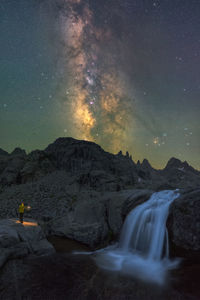 Anonymous traveler with torch contemplating waterfall streaming among rough rocky terrain under night starry sky with bright glowing milky way person