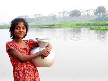 Portrait of young woman drinking water while standing against lake