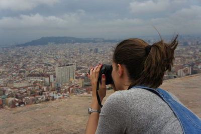 Woman photographing cityscape while standing on field against cloudy sky