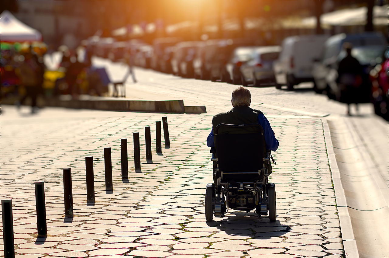 street, rear view, road, city, architecture, sunlight, adult, one person, transportation, footpath, men, city life, wheelchair, nature, mode of transportation, outdoors, infrastructure, sitting, full length, walking, vehicle, city street, medical equipment, differing abilities, travel, sidewalk, on the move
