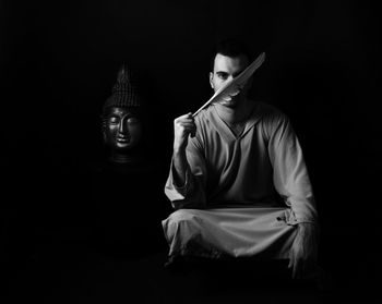 Portrait of man holding feather while sitting by buddha statue against black background