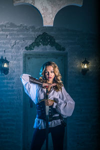 Portrait of woman in costume holding sword standing against door of abandoned building at night