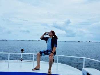 Full length of man drinking beer while sitting on boat at sea against sky