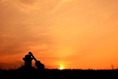 Silhouette guitar by bollard on field against orange sky