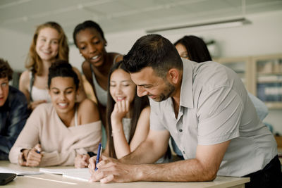 Smiling tutor teaching teenage students while leaning over table in classroom