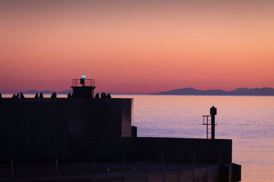 Silhouette building by sea against orange sky