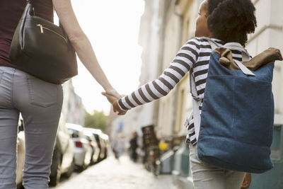 Midsection of woman holding hands with daughter while walking on sidewalk in city