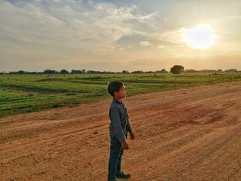 Side view of boy standing on field against sky during sunset
