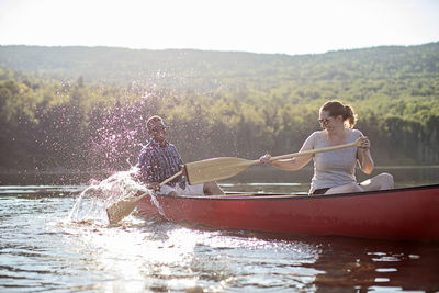 Couple splash each other with canoe paddles while boating on lake