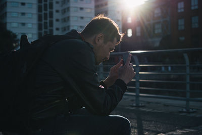 Serious man using smart phone while sitting by city street on sunny day