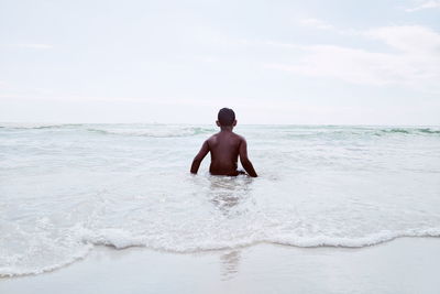 Rear view of shirtless boy standing in sea against sky