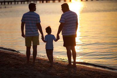 Rear view of men holding hands of boy while standing at beach during sunset