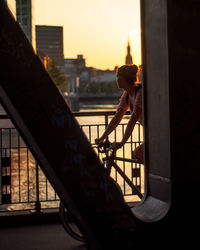 Woman riding bicycle on bridge in city during sunset