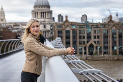 Portrait of smiling woman standing on bridge over river against buildings