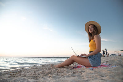 Side view of woman sitting at beach against sky