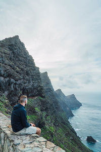 Rear view of man sitting on rock by sea against sky