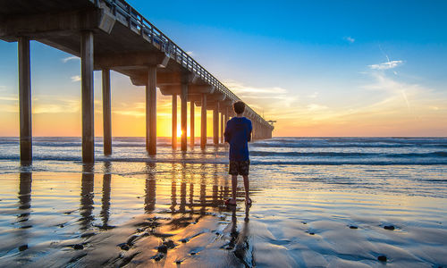 Rear view of man walking by sea against sky during sunset