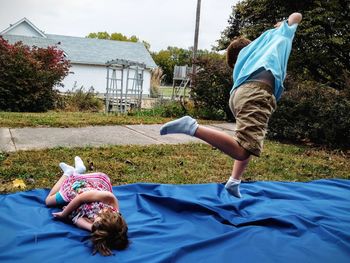 Siblings playing on blue fabric in yard by house against sky