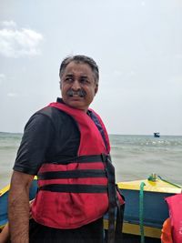 Man wearing life jacket looking away while standing on rowboat in sea against sky