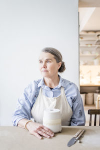 Senior female potter looking away while sitting at table with vase against white wall in workshop