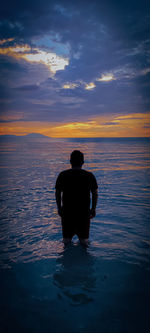 Rear view of man standing in sea against sky during sunset