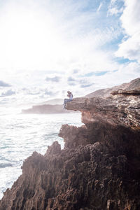 Woman sitting on cliff by sea against cloudy sky during sunny day