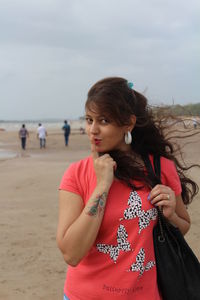 Portrait of woman with finger on lips at beach against cloudy sky