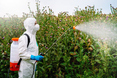 Person in protective workwear and mask holding sprinkler and spraying herbicide on land