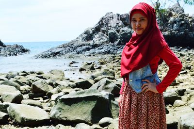 Portrait of young woman standing on rock at beach against sky