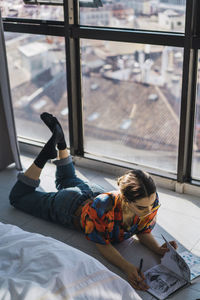 High angle view of young woman with book lying on tiled floor at home