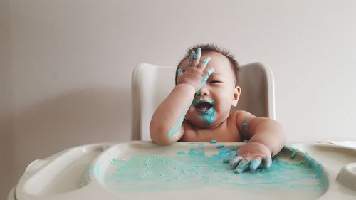 Playful baby playing with paint while sitting on chair at table against wall