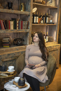 Thoughtful pregnant woman sitting by shelf on chair