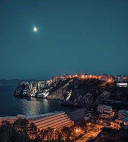 Illuminated buildings by sea against clear sky at night