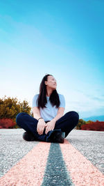 Young woman sitting on highway against blue sky