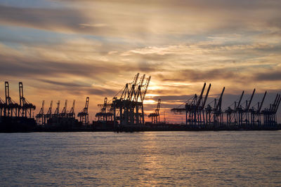 Silhouette cranes at harbor against sky during sunset