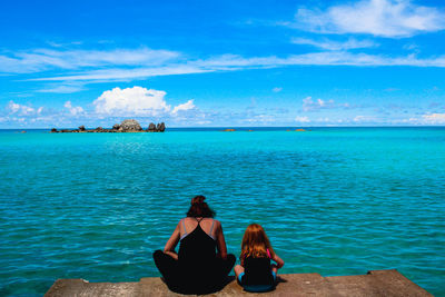 Rear view of mother and daughter sitting by sea against blue sky