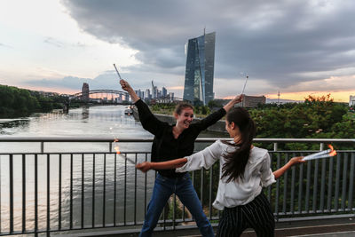 Happy playful friends by railing on bridge in city against sky