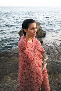 Contemplative woman wrapped in blanket standing on shore