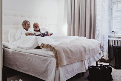 Senior woman reading newspaper while having coffee by man relaxing on bed in hotel