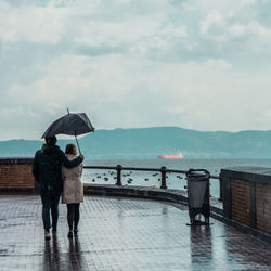 Rear view of couple with umbrella walking on wet road against sky during rainfall