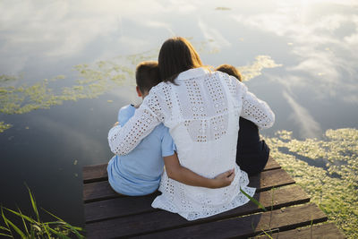 Mother embracing sons sitting on jetty in front of lake